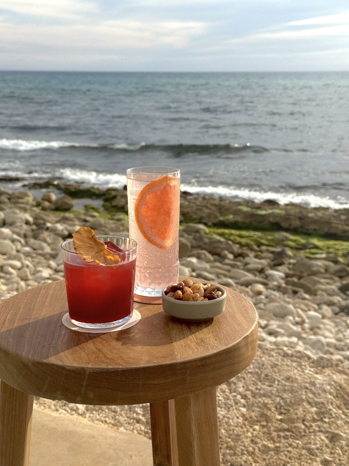 Two cocktails and a bowl of mixed nuts on a wooden table by the rocky Batroun shoreline.